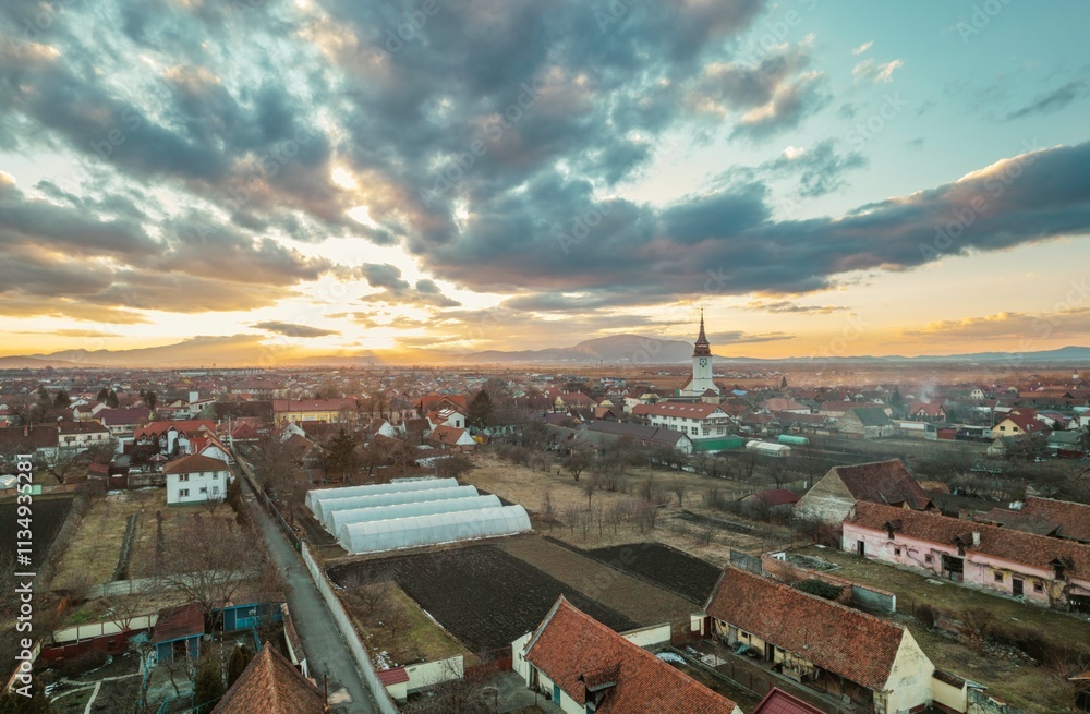 Obraz premium Captivating sunset envelops Rasnov with vibrant colors illuminating the rooftops. The nearby mountains add a majestic backdrop to this serene landscape, showcasing nature's beauty