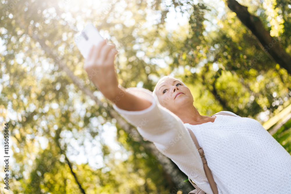 Low-angle view of stylish gray-haired senior woman taking selfie using mobile phone during walking in city park, on sunny summer day. Content elderly female taking selfie surrounded by nature. Low-angle view of stylish gray-haired senior woman taking selfie using mobile phone during walking in city park, on sunny summer day. Content elderly female taking selfie surrounded by nature.