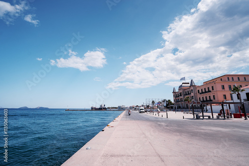 Fototapeta Naklejka Na Ścianę i Meble -  Travel by Greece. A scenic coastal promenade with a clear blue sky, calm sea, and historic buildings. The area is peaceful with few people and a distant view of islands on the horizon.