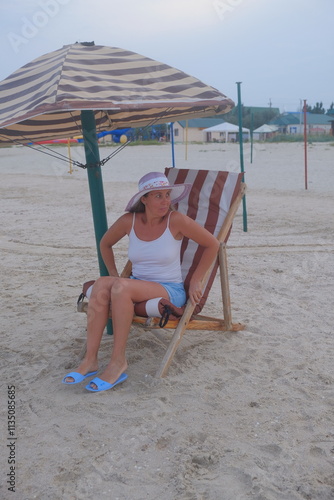 child on the beach woman sitting in a lounge chair on the seashore with a hat in summer autumn spring and holding her hand on a hat panama european appearance there are emotions on her face in shorts  photo