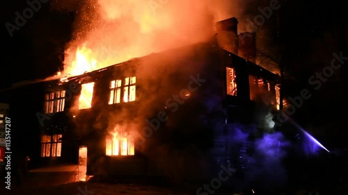 Firefighters are fighting a burning house fire. A fireman is watering the flames in the house at night. The fiery disaster and the work of rescuers. A fire in the Building.