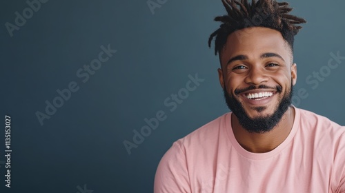 A young, bearded man with braided hair and a wide smile wears a pink shirt, radiating happiness and warmth in front of a plain background.