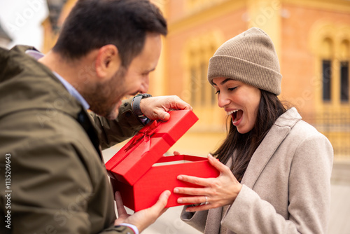 A happy Caucasian couple shares gifts on International Women's Day. The woman, dressed in a stylish coat and beanie, receives a present from the man in a casual jacket. photo