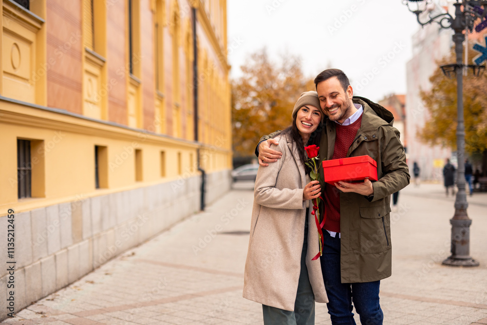Fototapeta premium Mid adult Caucasian male surprises wife with a present and red rose. They enjoy a romantic day outdoors. She wears a cozy coat and beanie, while he sports a jacket.