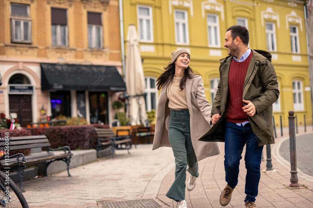 Obraz premium Smiling Caucasian couple enjoys a fun run on the street. Man in a green jacket and red sweater, woman in a beige coat and hat.