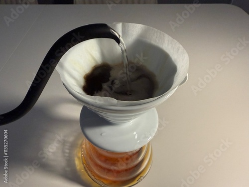 A minimalist pour-over coffee setup featuring a ceramic dripper, paper filter, glass carafe, and a black gooseneck kettle on a white table. photo