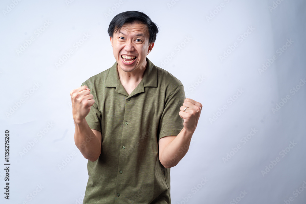 An Asian man in a casual green shirt clenches his fists and grins with excitement, standing against a clean white background. The image conveys joy, success, or celebration. An Asian man in a casual green shirt clenches his fists and grins with excitement, standing against a clean white background. The image conveys joy, success, or celebration.