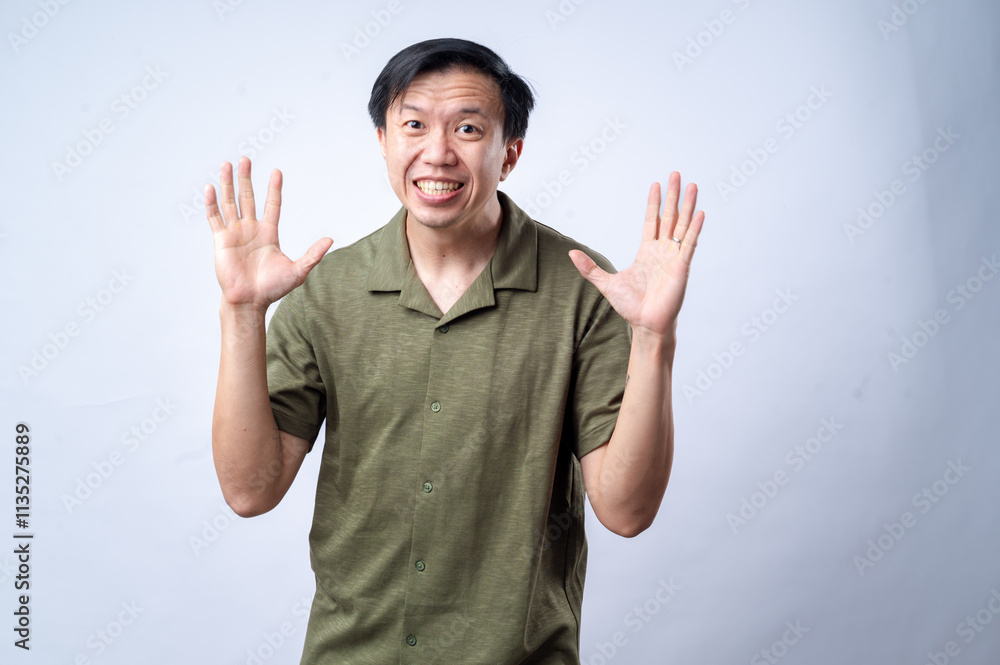 Fototapeta premium An Asian man in a casual green shirt raises his hands and shouts with an exaggerated expression, standing against a clean white background. The image conveys excitement, surprise, or dramatic reaction