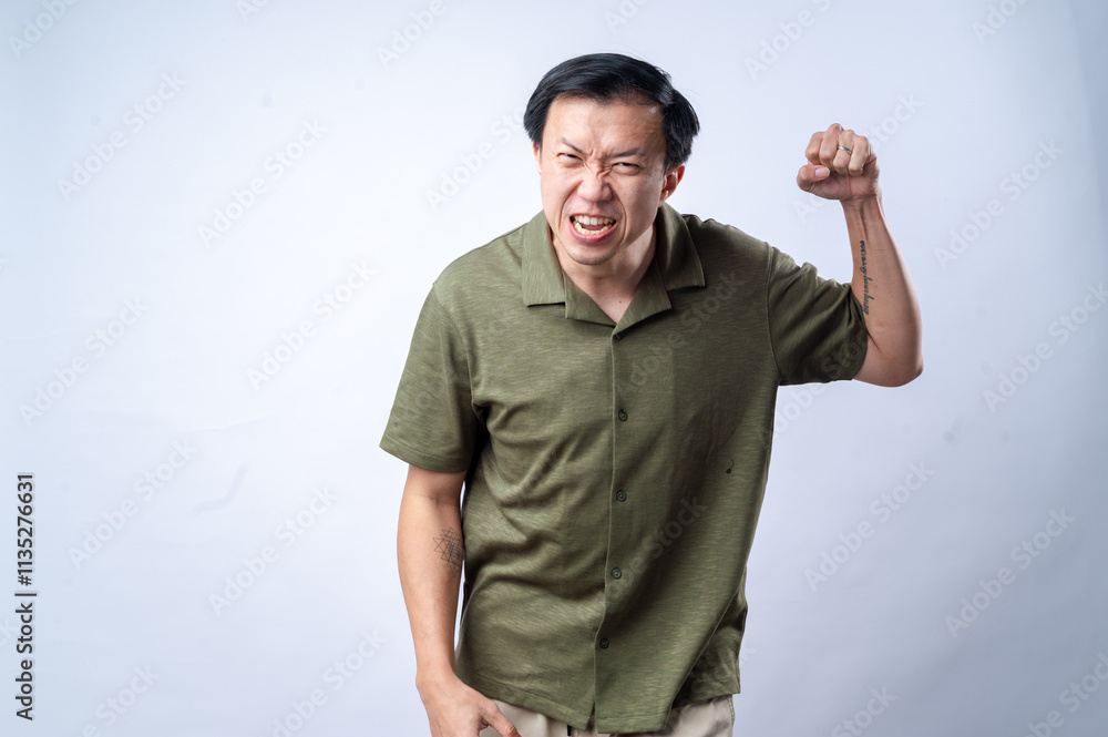 Naklejka premium An Asian man in a green shirt shows anger and determination with a clenched fist raised and a fierce expression, conveying a sense of defiance or frustration, against a plain white background