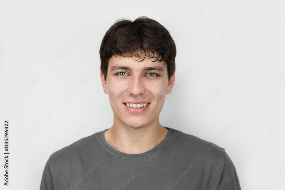 Portrait of a young man dressed in a gray shirt, smiling warmly against light background. The guy has dark curly hair and white healthy teeth