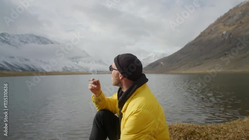 Wallpaper Mural Handsome Man Traveler in a yellow jacket stands by a beautiful mountain lake and smokes a cigarette, reflecting on nature's scenic beauty Torontodigital.ca
