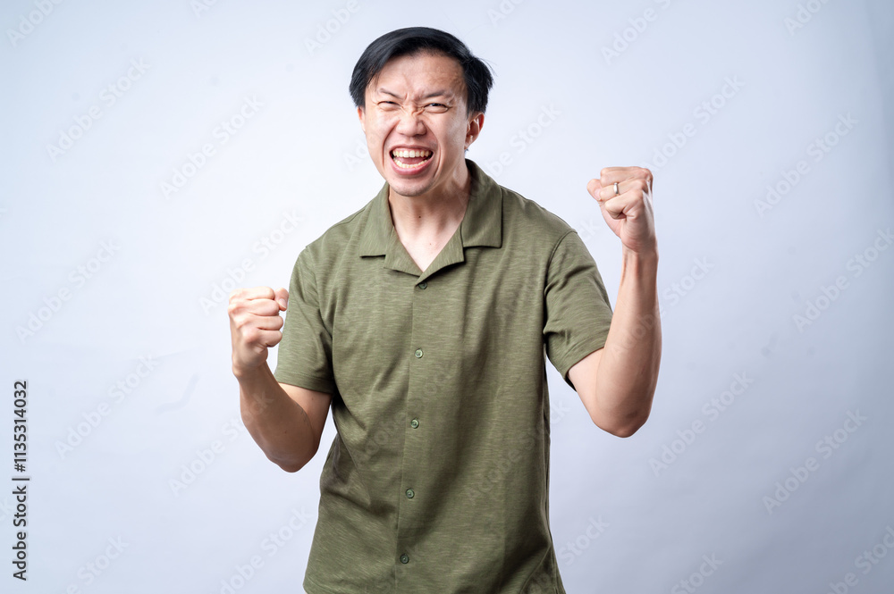 An Asian man in a casual green shirt clenches his fists and grins with excitement, standing against a clean white background. The image conveys joy, success, or celebration. An Asian man in a casual green shirt clenches his fists and grins with excitement, standing against a clean white background. The image conveys joy, success, or celebration.