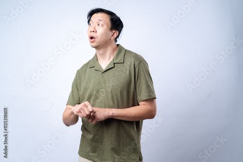 An Asian man in a casual green shirt looks to the side with a thoughtful expression, clasping his hands together, standing against a clean white background. The image portrays contemplation photo