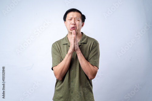 Fotografie An Asian man in a green shirt with a pleading expression, hands clasped together as if begging or praying, looking upward, standing against a plain white background in a studio setting
