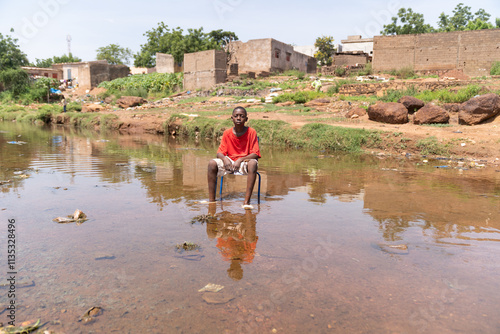 Young African boy sitting on a chair in the middle of a flooded area symbolizing the impact of climate change on life in poor developing countries