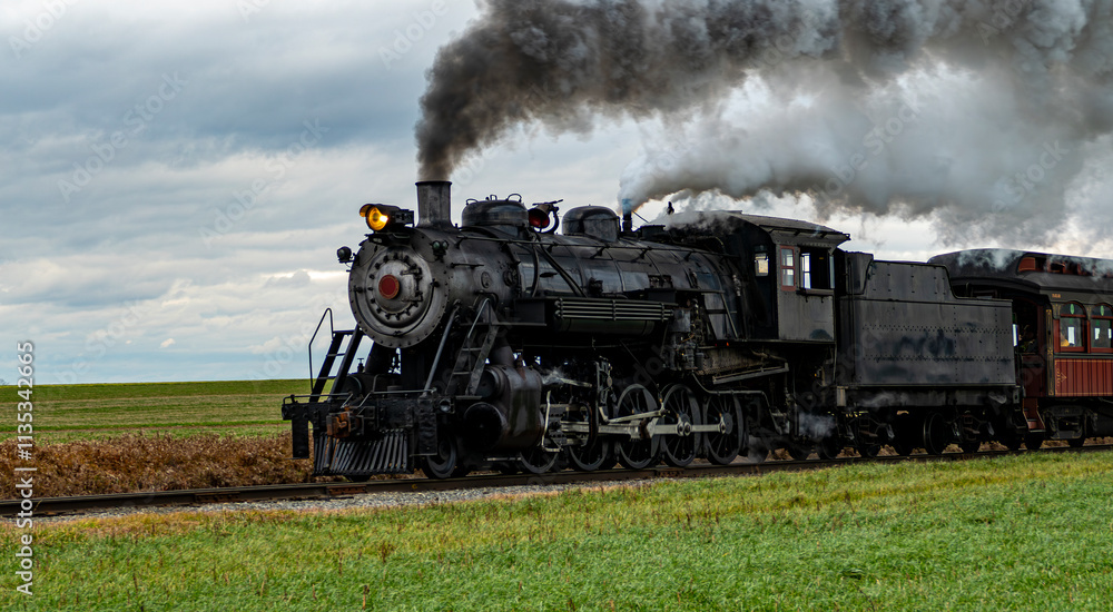 Fototapeta premium A vintage steam train chugs along railway tracks in a vast rural area, emitting thick plumes of smoke against a backdrop of dark clouds. Nearby fields exhibit golden crops.