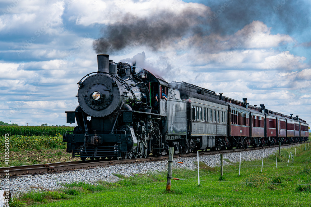 Naklejka premium A vintage steam train puffs black smoke as it moves along the railway, surrounded by lush green cornfields and under a bright blue sky with fluffy clouds, creating a picturesque countryside view.