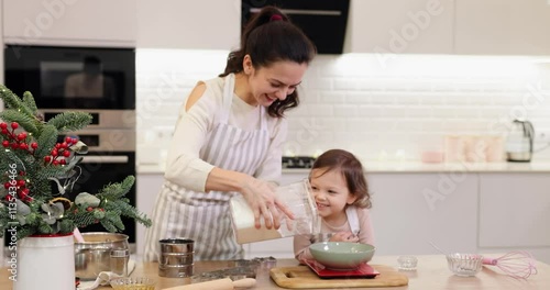 smiling mother and her cute little child daughter in aprons preparing the dough in the kitchen, mom and kid weigh flour on scales, Christmas time