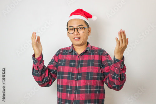 An Asian man in a red plaid shirt, and a Santa hat expresses surprise with open arms and wide eyes. photo