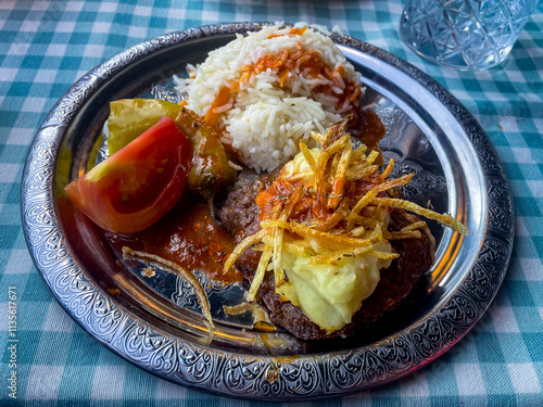 Istanbul, TR - Oct 23, 2024 A plate of Hasanpaşa meatballs topped with mashed potatoes and crispy fries, served with rice, grape leaves, tomato, and flavorful sauce. A Turkish delight!