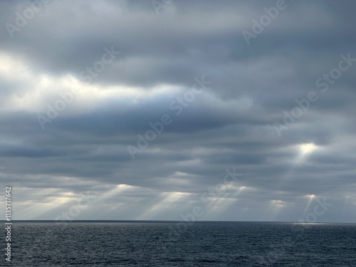 Dramatic sky with dark clouds and beams of sunlight piercing through, casting light on the calm ocean below. photo