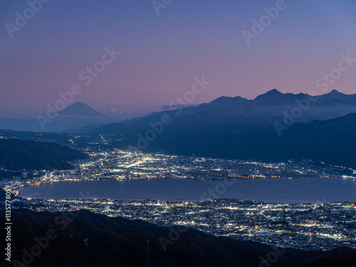 高原の展望台より山と湖と街並みを望む、夕焼けの空で少し霞が掛かり水平線はピンク色に染まり山々はシルエット、薄暗くなり街明かりが宝石箱
の様に綺麗に輝いています、空の比率が多くて左側に山、中央に街明かりと湖がある横構図です。 photo