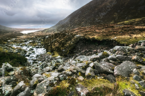 A view of a valley from the bottom of a rocky slope. The valley is filled with rocks and grass, with a stream running through it. The sky is overcast, with the sun partially covered by clouds. photo