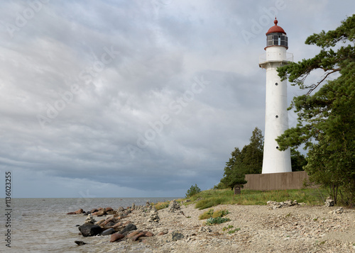 A white lighthouse, rocky shoreline, surrounded by lush greenery. The cloudy sky looms overhead, adding a sense of drama to the serene coastal scene.  Estonian island. Sea, rocky beach, summer evening