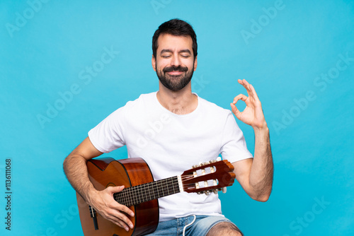 Young man with guitar over isolated blue background in zen pose