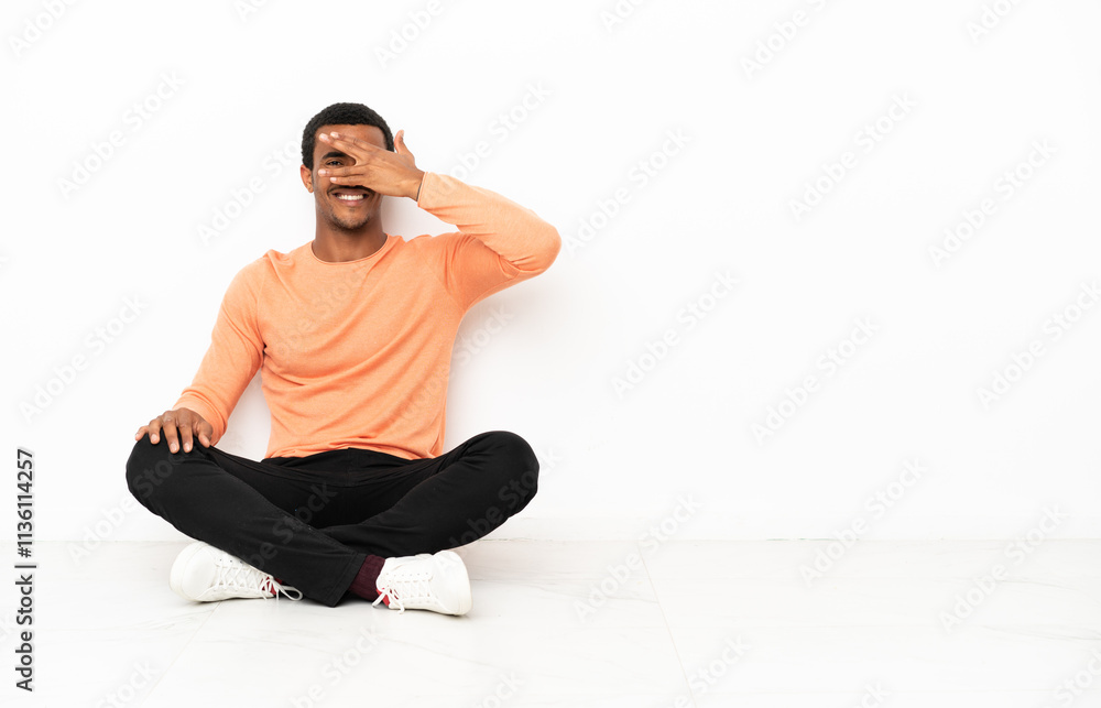 Fototapeta premium African American man sitting on the floor over isolated copyspace background covering eyes by hands and smiling
