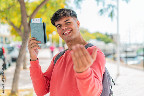 Young hispanic man at outdoors holding a passport and doing coming gesture photo