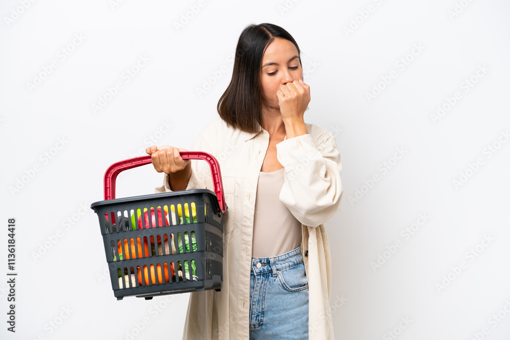 Young woman holding a shopping basket full of food isolated on white background having doubts Young woman holding a shopping basket full of food isolated on white background having doubts