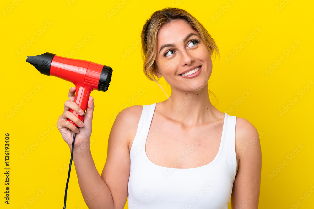 Young Uruguayan woman holding a hairdryer isolate don yellow background looking up while smiling Young Uruguayan woman holding a hairdryer isolate don yellow background looking up while smiling