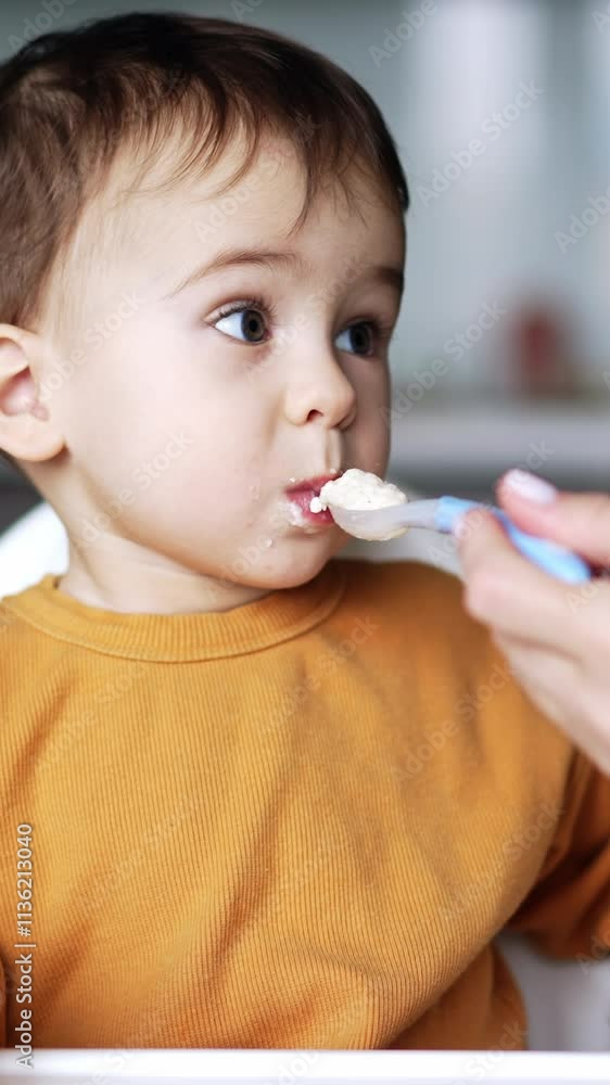 Beautiful Caucasian baby boy having dinner at home. Child takes a spoonful from mom and sits still with a full mouth looking straight into camera thoughtfully. Vertical video Beautiful Caucasian baby boy having dinner at home. Child takes a spoonful from mom and sits still with a full mouth looking straight into camera thoughtfully. Vertical video