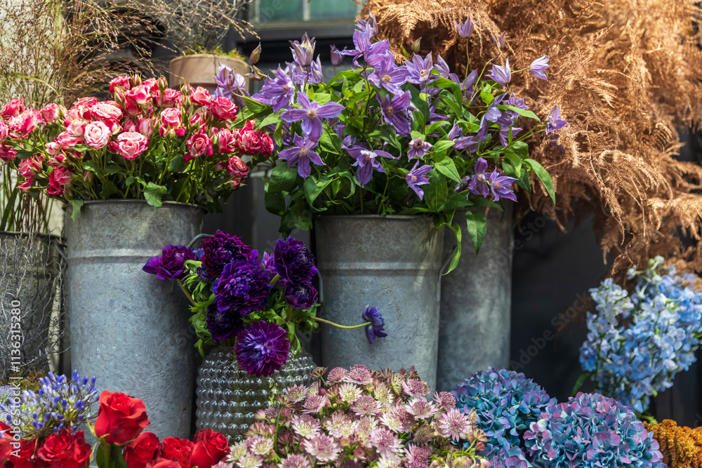 Fototapeta premium Purple clematis, orange roses, windflowers, anemones, hydrangeas, asparagus, and astrantia displayed in vases and tin buckets for sale, ideal for enhancing interior decor.