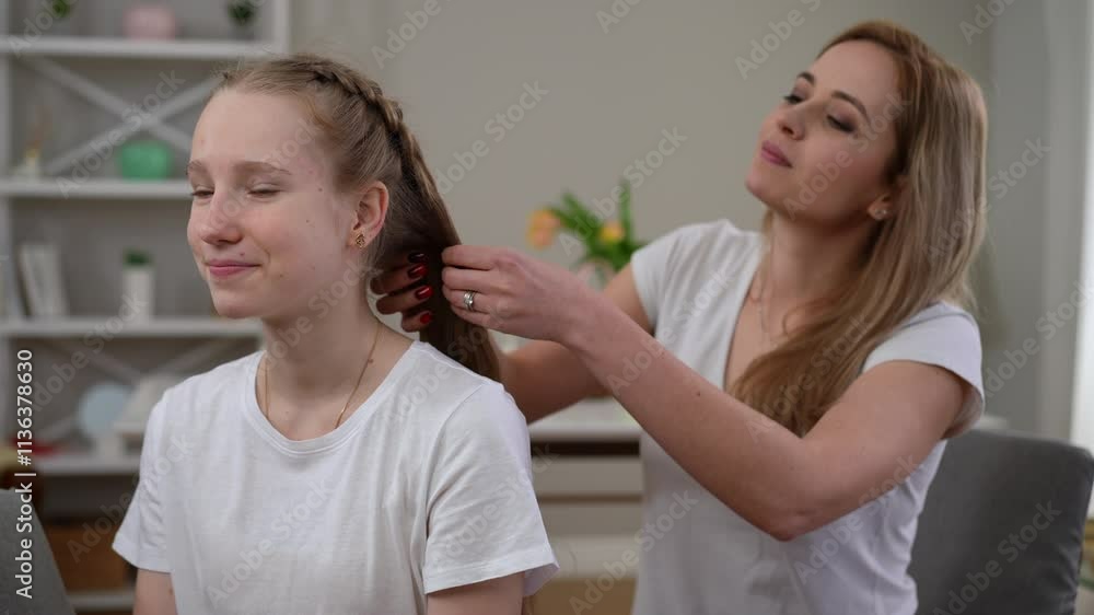 A mother gently braids her teenage daughter's long hair as they sit together in a cozy room. The teenager smiles softly, enjoying the care and attention. A vase of flowers and a bookshelf add warmth A mother gently braids her teenage daughter's long hair as they sit together in a cozy room. The teenager smiles softly, enjoying the care and attention. A vase of flowers and a bookshelf add warmth