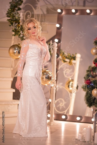 Obraz na płótnie glamorous blonde in a white shiny dress with white hair stands at a large mirror, next to a decorated Christmas tree . w salonie