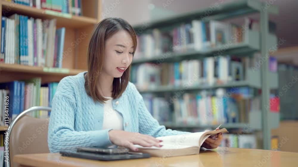 A woman is sitting at a table in a library, reading a book. She is smiling and she is enjoying her time. The library is filled with bookshelves, and there are several books scattered around the room A woman is sitting at a table in a library, reading a book. She is smiling and she is enjoying her time. The library is filled with bookshelves, and there are several books scattered around the room