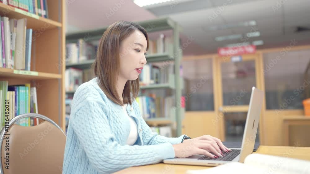 A woman is sitting at a desk in a library, typing on a laptop. Concept of focus and concentration, as the woman is likely working on a project or studying. The library setting suggests a quiet A woman is sitting at a desk in a library, typing on a laptop. Concept of focus and concentration, as the woman is likely working on a project or studying. The library setting suggests a quiet