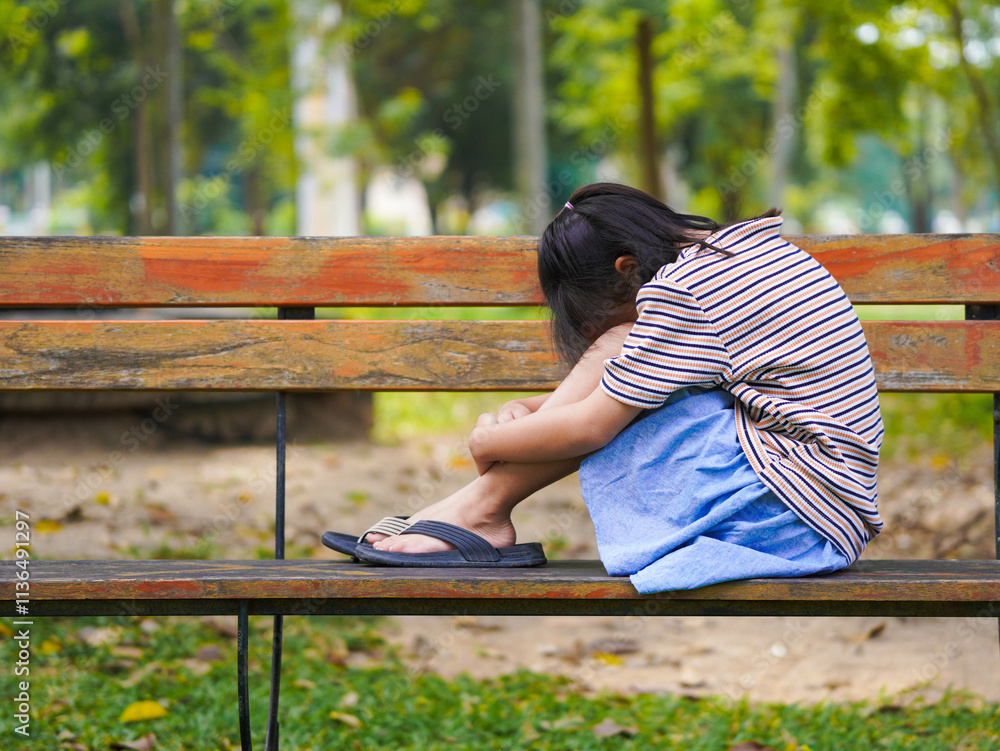 Naklejka premium A sad little girl is sitting alone on bench in park. The little girl is unhappy and anxious because she has no one to play with.