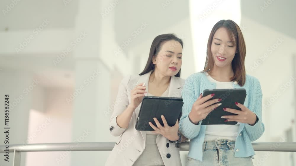 Two women are looking at their tablets, one of which is a tablet computer. Scene is casual and relaxed, as the women are sitting on a railing and using their devices Two women are looking at their tablets, one of which is a tablet computer. Scene is casual and relaxed, as the women are sitting on a railing and using their devices