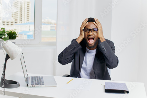 Stressed businessman in formal suit sitting at desk holding head in hands with laptop and documents nearby photo