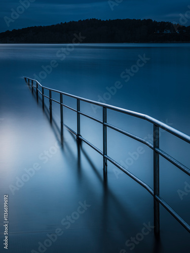 Calm water reflecting twilight with a metal railing leading into the lake at dusk photo