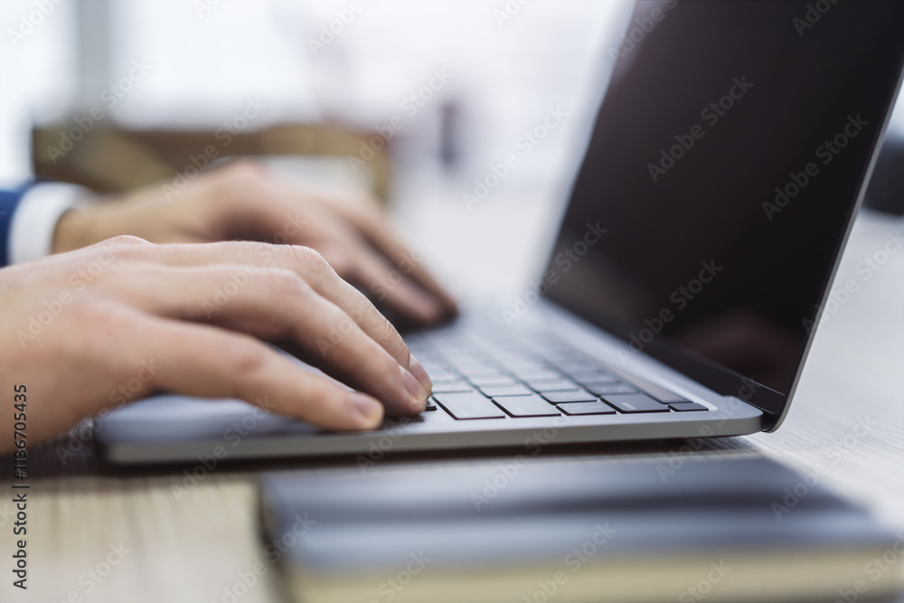 The photo presents a close-up of a man's hands as they type on a cutting-edge laptop keyboard, against a blurred office backdrop The photo presents a close-up of a man's hands as they type on a cutting-edge laptop keyboard, against a blurred office backdrop