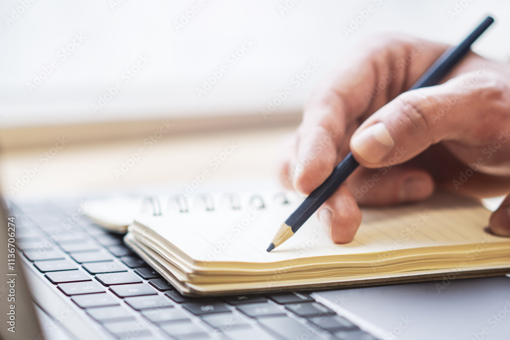 Close-up depiction of a man's hand taking notes in a notepad on a sleek laptop, with a blurred background Close-up depiction of a man's hand taking notes in a notepad on a sleek laptop, with a blurred background