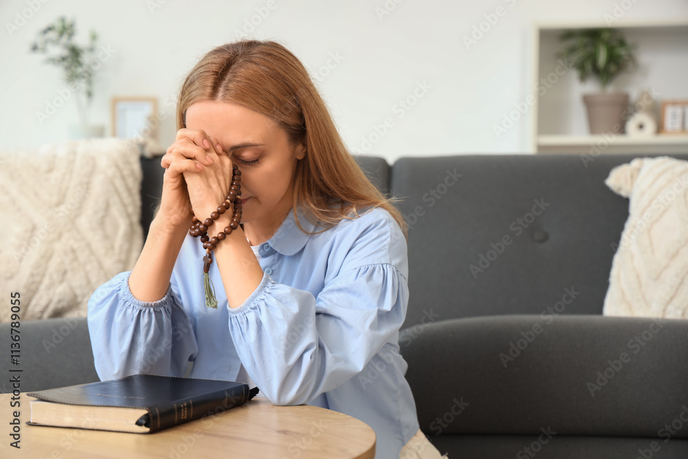 Mature woman praying with beads and Holy Bible on table at home