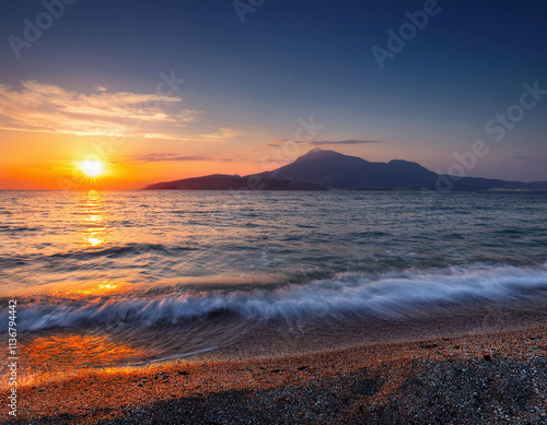 The sun sets dramatically over a mountain silhouette, casting golden reflections on the waves crashing onto a sandy beach. photo