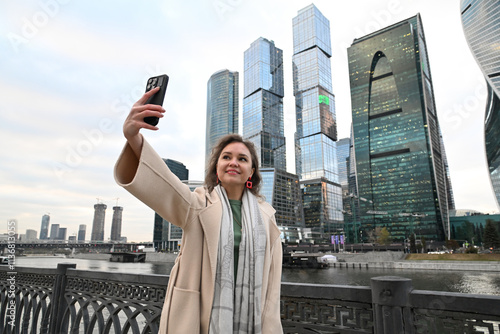 A young woman of European appearance takes a selfie with a smartphone against the backdrop of the Moscow City skyscrapers and the embankment outdoors