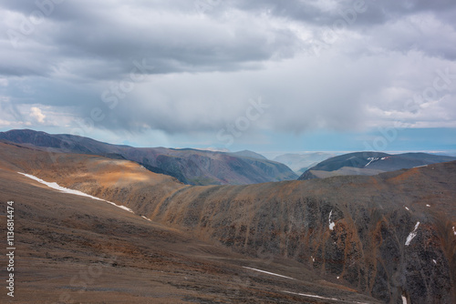 Dramatic layered top view from pass above large sharp ridge to multicolor alpine deep valley among big cliffs far away in rain under cloudy sky. High rocky mountains on horizon under rainy clouds.