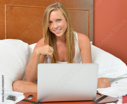 Sexy blonde woman in bed using laptop computer with tv remote and journal nearby